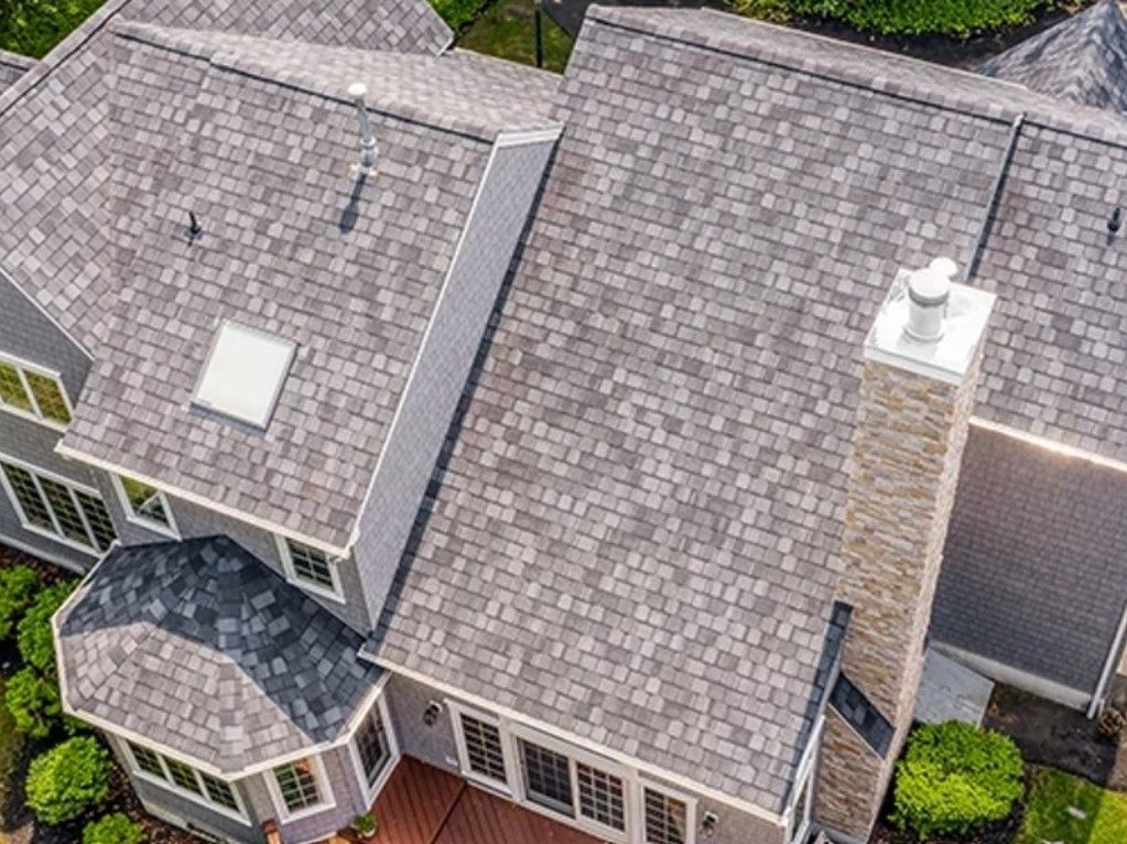 An aerial view of a multi-gabled house with a light gray shingled roof, a stone chimney, and a small deck.