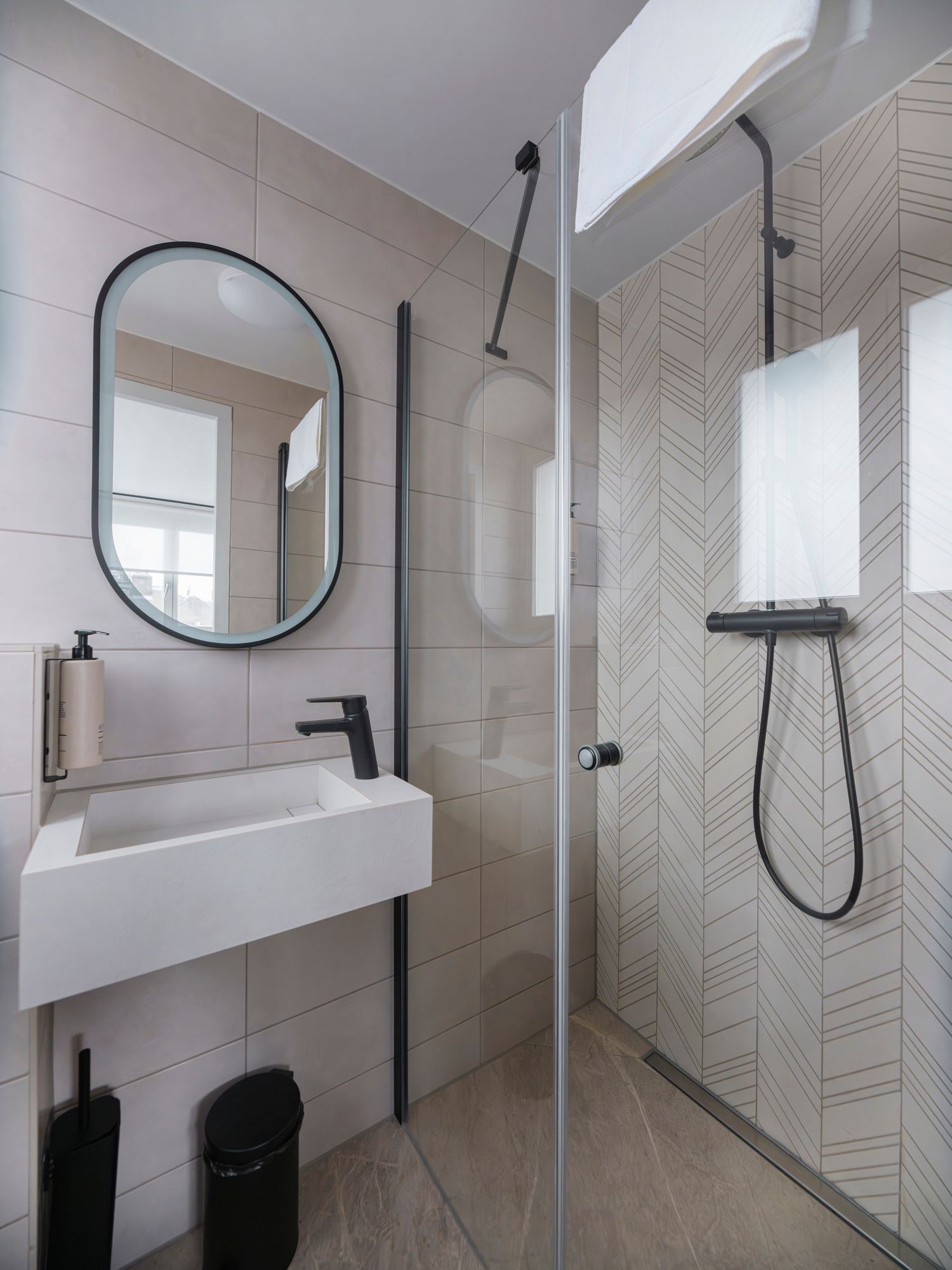 Modern bathroom with a wall-mounted white sink, oval mirror, and a glass-enclosed shower with patterned tile walls.