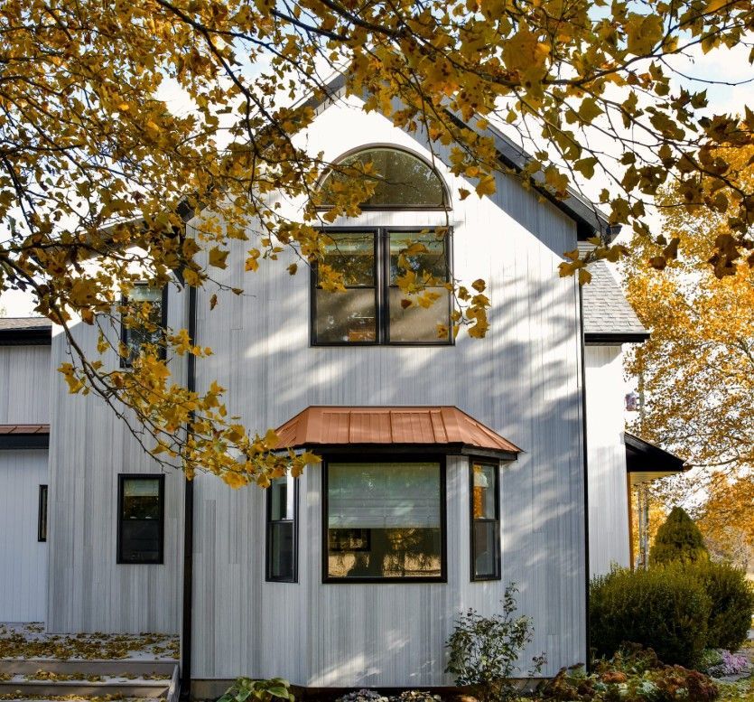 A two-story house with gray vertical siding, a copper-roofed bay window, and dark frames, framed by yellow autumn leaves.