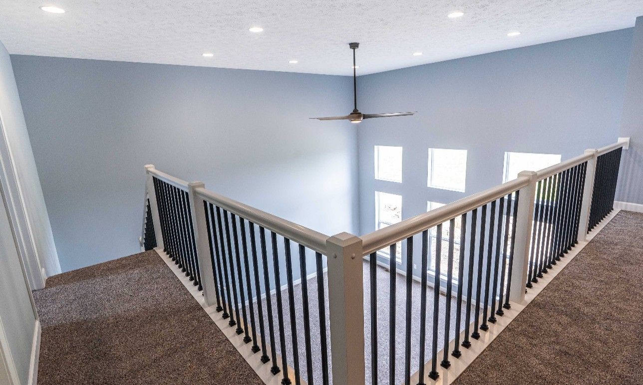 A high-angle view from a carpeted loft looking down into a living room with light blue walls, high windows, and a ceiling fan.