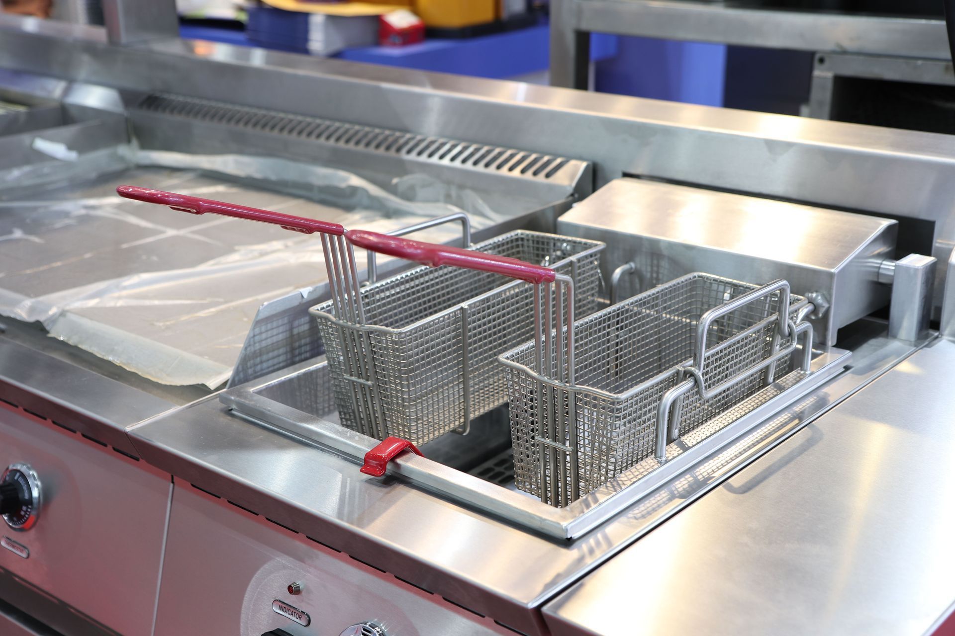 Stainless steel commercial deep fryer with two wire baskets, a drainboard, and red-handled tongs.