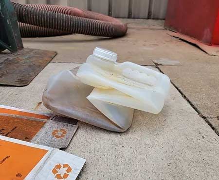A crushed plastic jug and recycling materials sit on a concrete surface, with a hose and metal piece in the background.