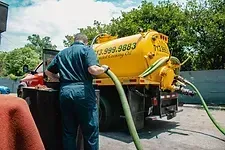Man operating a yellow septic tank truck, holding a green hose outdoors.