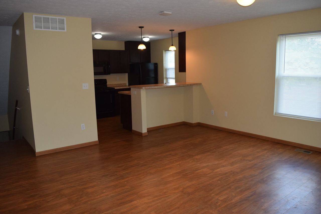 An empty living room with hardwood floors and a kitchen in the background.