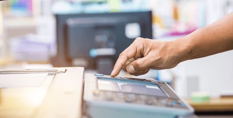 Person's hand presses a button on a copier, likely to start a print job, in an office setting.