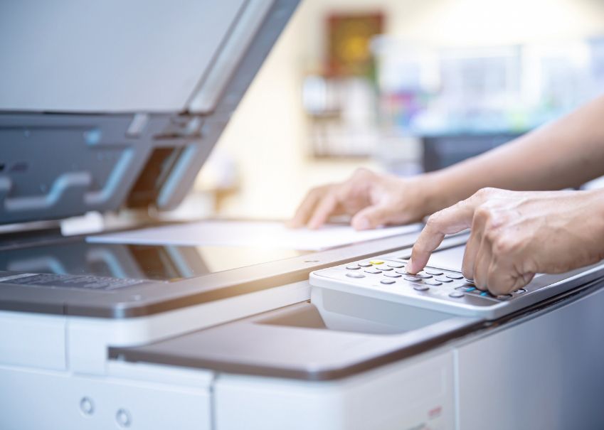Person using a copier, pressing buttons on the control panel. Office setting with sunlight.