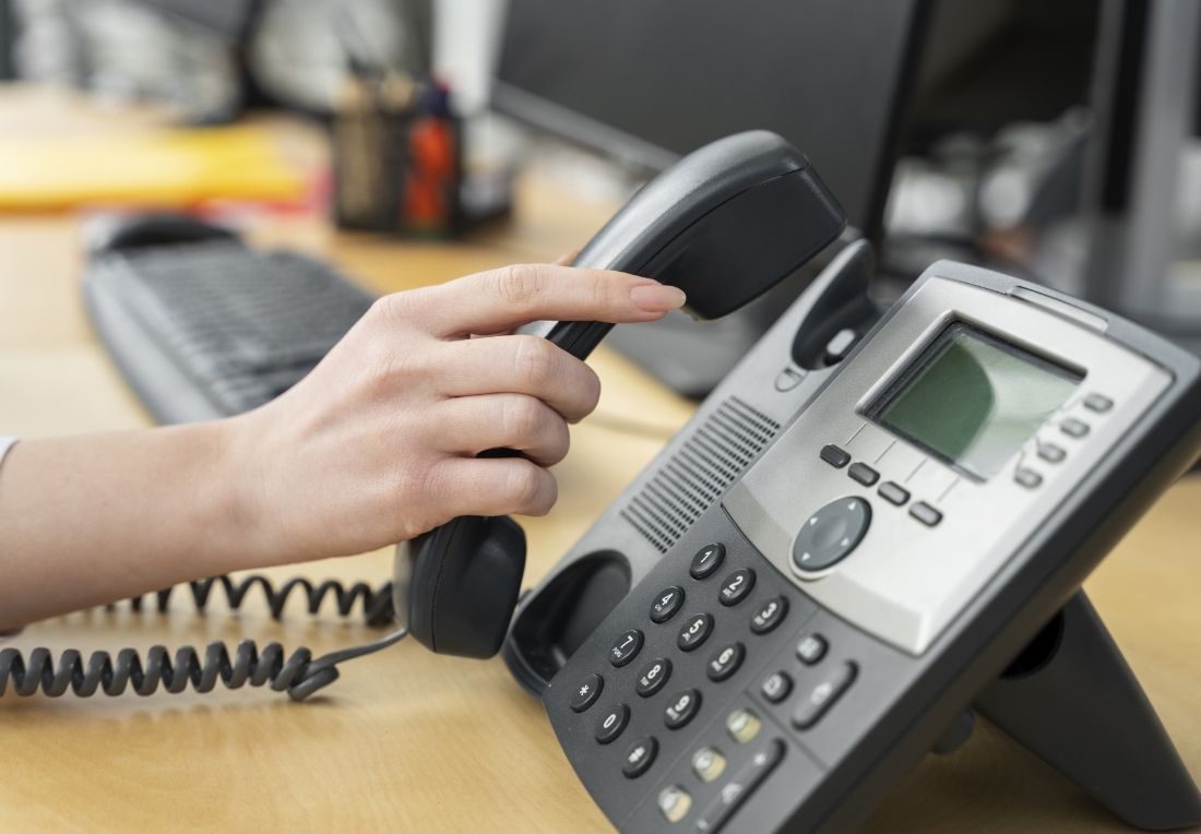 Hand lifting a black telephone receiver, office desk in background.