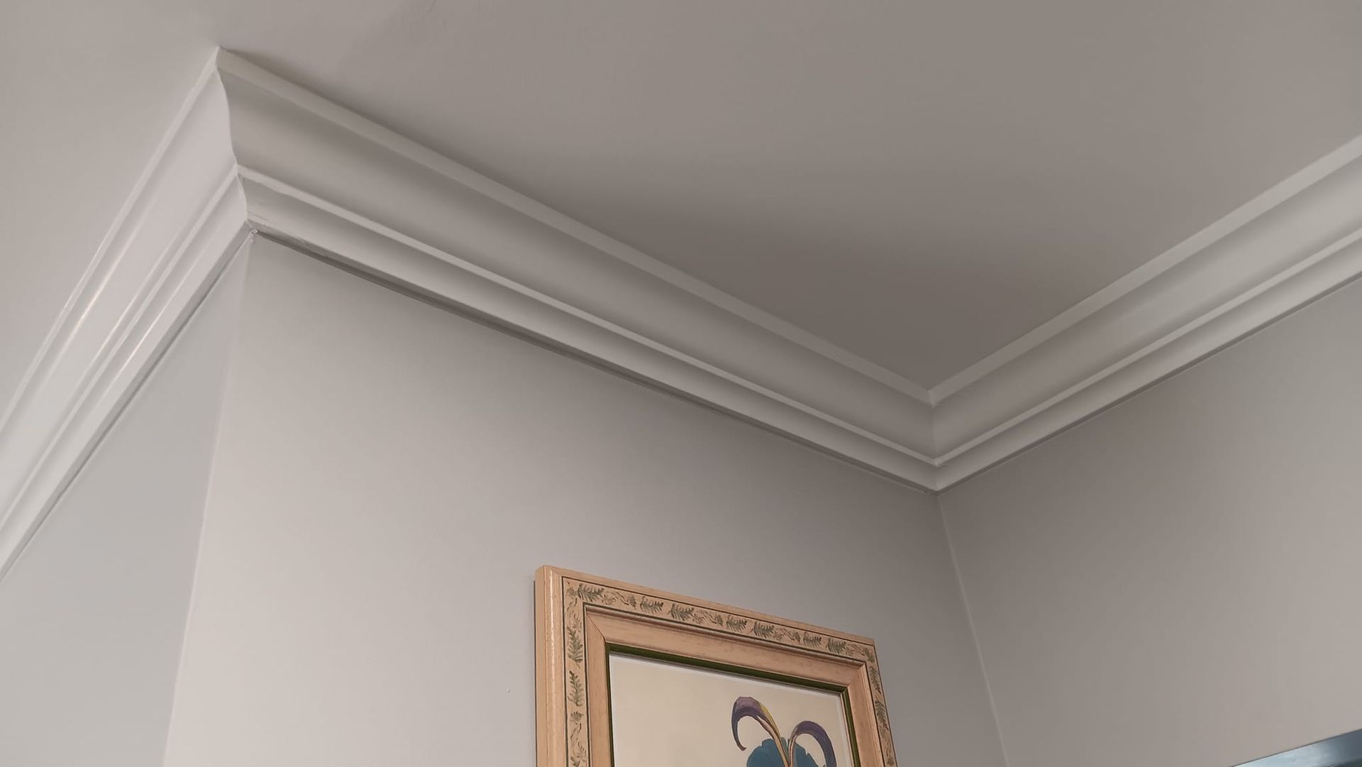 Corner of a room with white crown molding. A framed picture hangs below the molding. Gray walls.