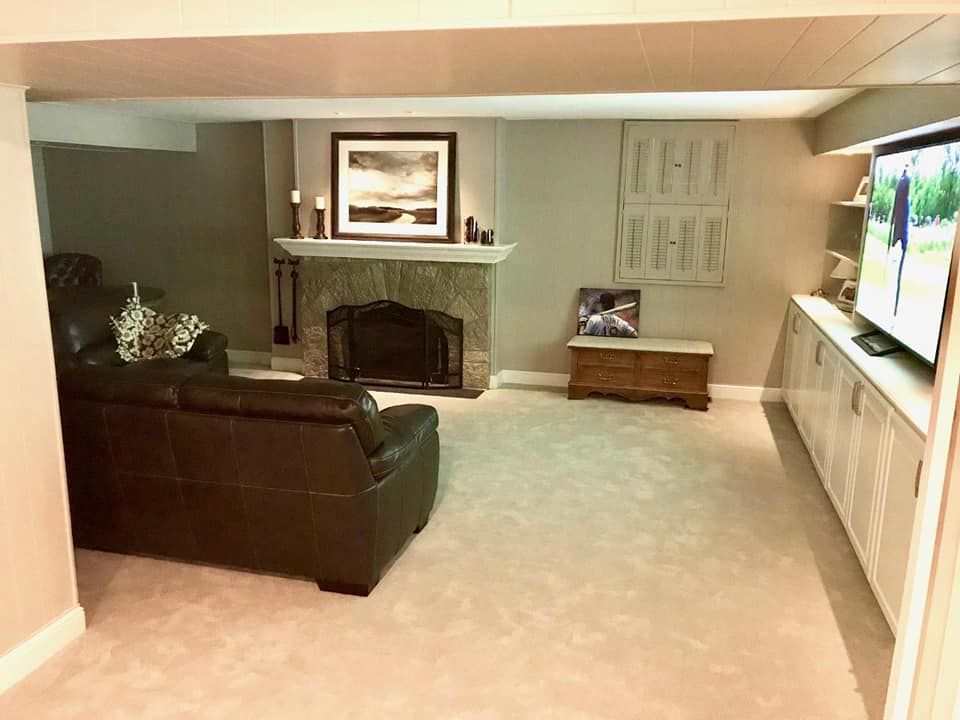 Cozy basement living room: brown leather couch, stone fireplace, TV on a built-in cabinet, beige carpet, and neutral walls.