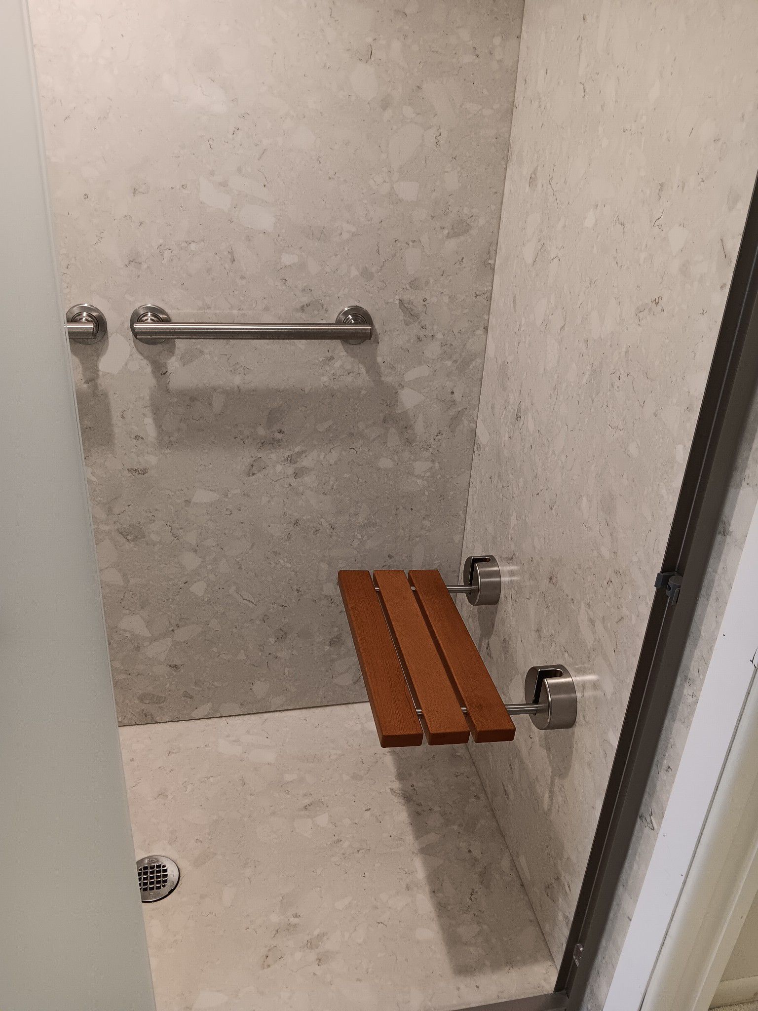 Shower stall with a wooden fold-down seat and a towel bar, set against gray speckled walls.