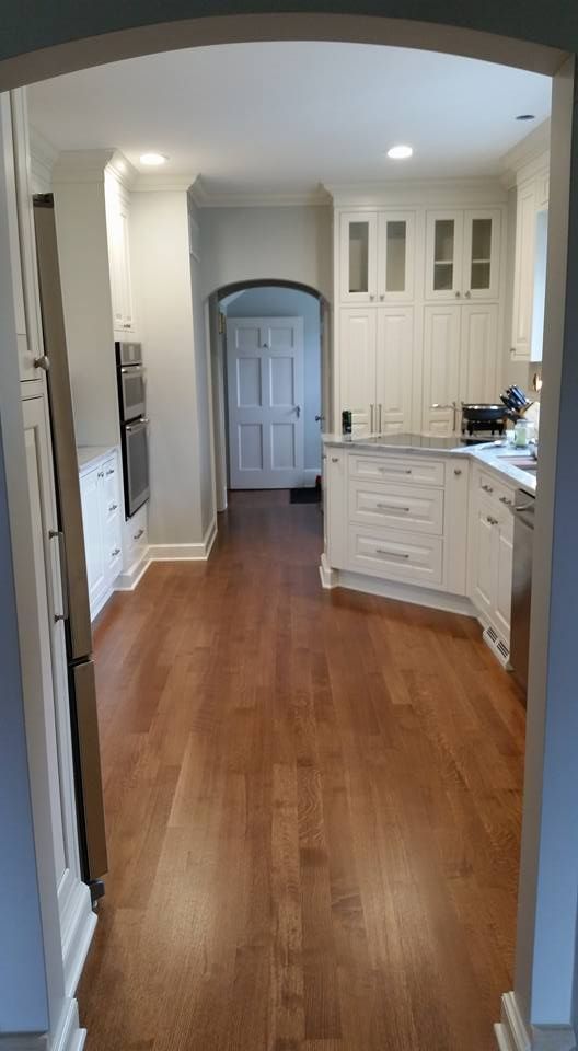 Interior view of a white kitchen with wood floors, cabinets, and appliances. An arched doorway is in the foreground.