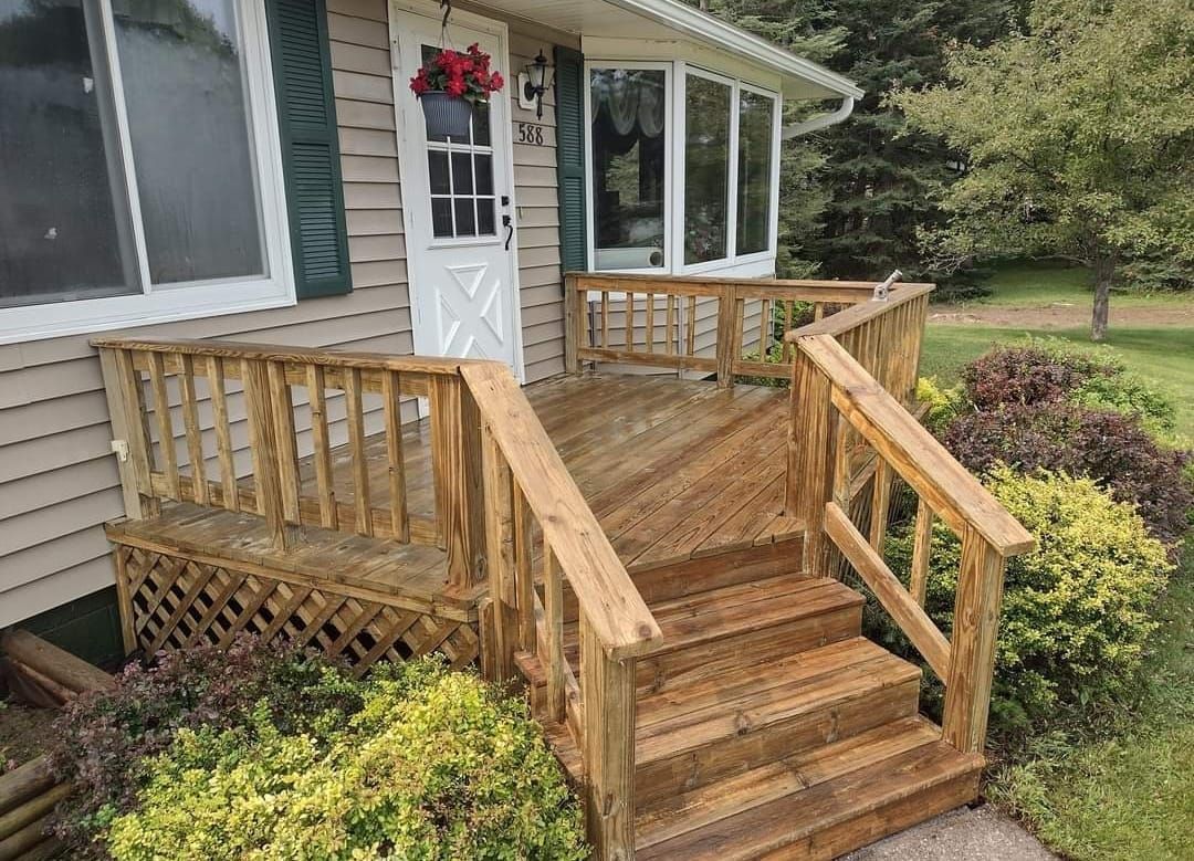 Wooden porch with stairs leading to a house. Steps and railing stained wood. Bushes and grass surrounding.