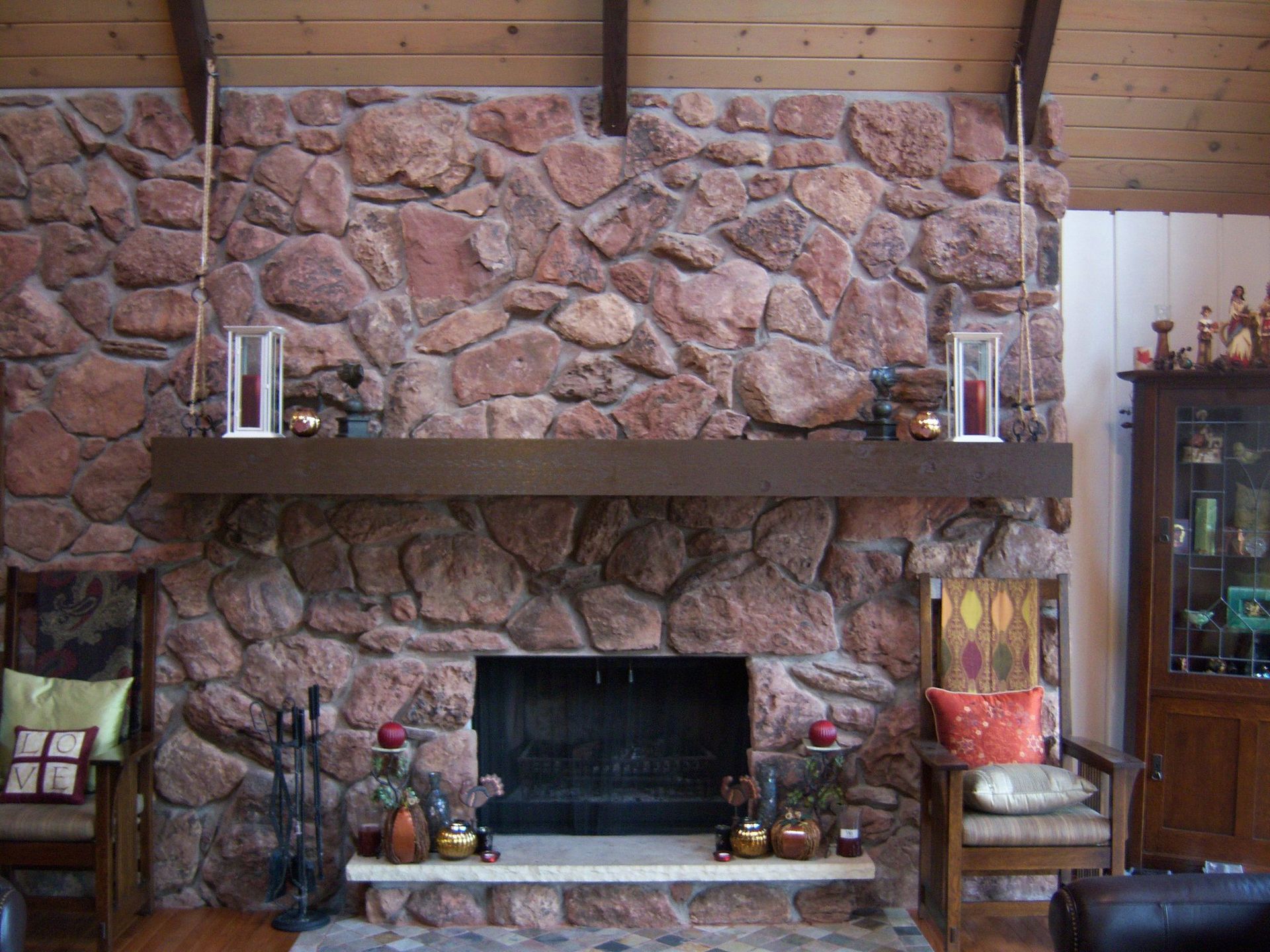 Stone fireplace with wooden mantel, adorned with candles and decorations; wooden beams, chairs.