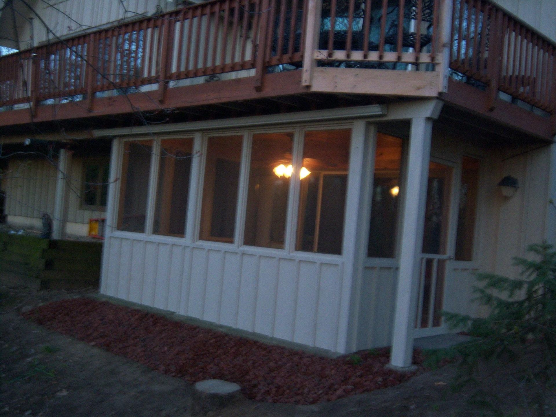 Screened-in porch beneath a wooden deck, painted white with red mulch and trees nearby.