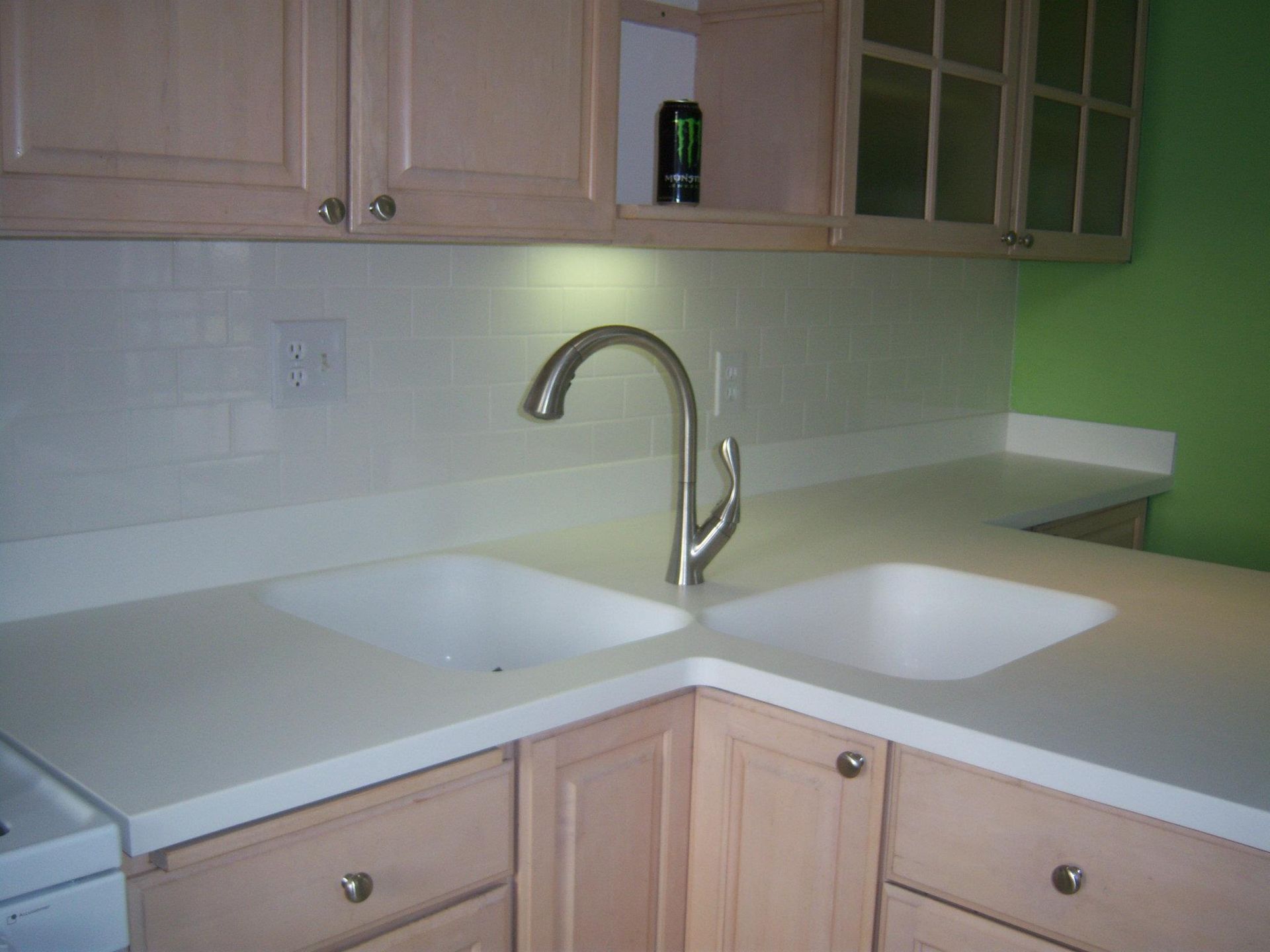 Kitchen sink with light-colored countertops, backsplash, and cabinets. A faucet is centered above the double sink. Green wall visible.