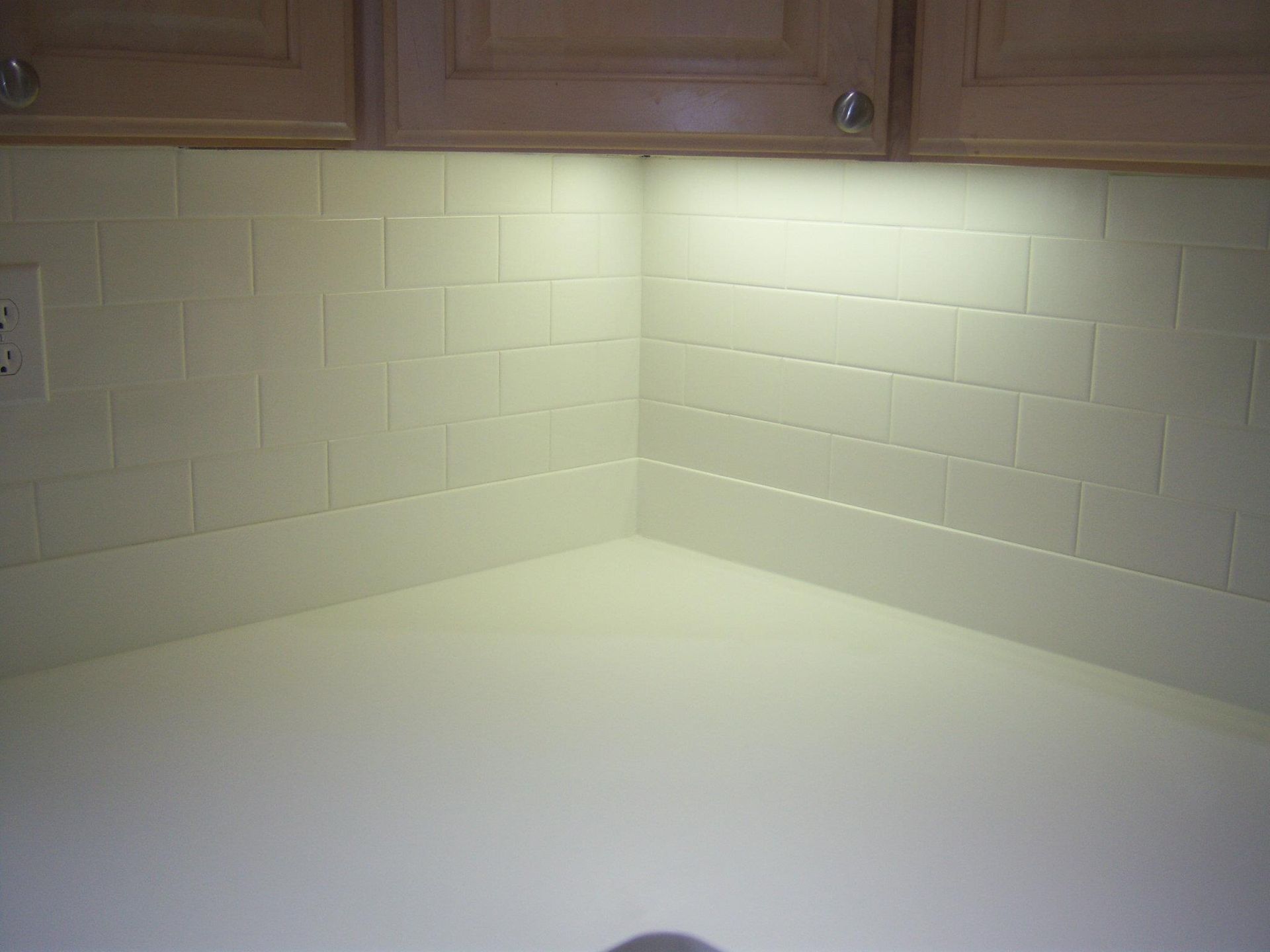 Kitchen corner with off-white tile backsplash, light-colored cabinets, and countertops.  Under-cabinet lighting.