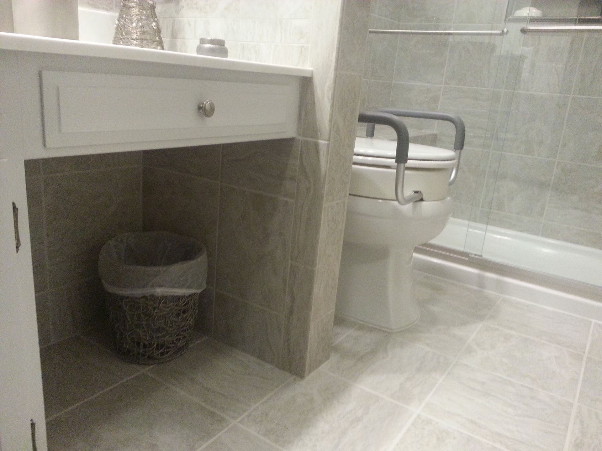 Bathroom with a raised toilet, grab bars, and a wastebasket under a cabinet.