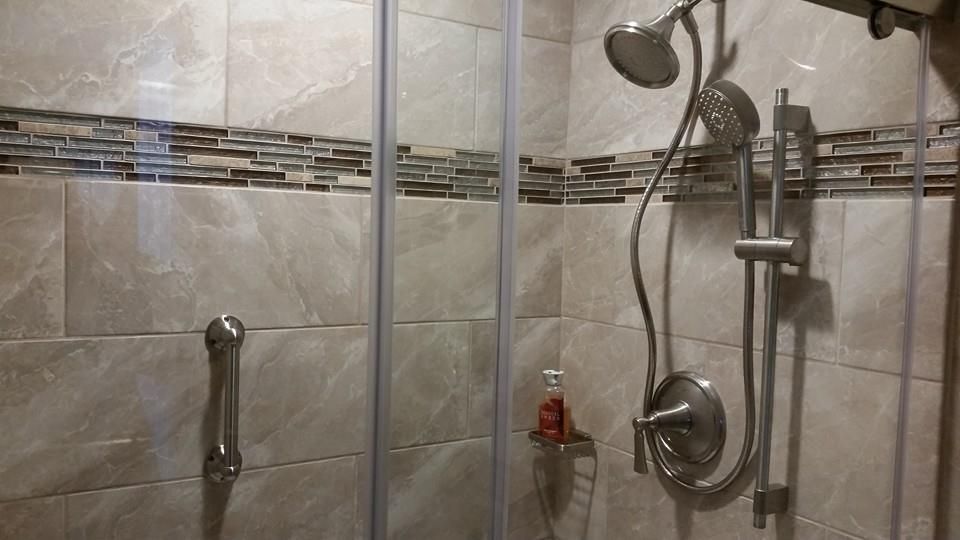 Shower with glass doors, beige tile, grab bar, showerhead, and a small shelf.
