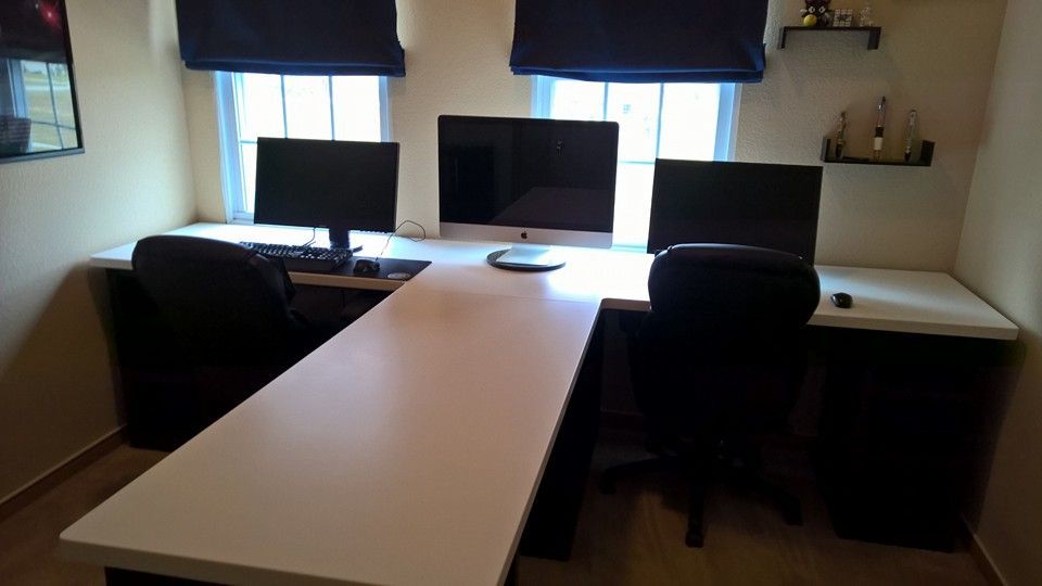 L-shaped desk with three computer monitors, two chairs, and two windows with blue blinds in a corner room.