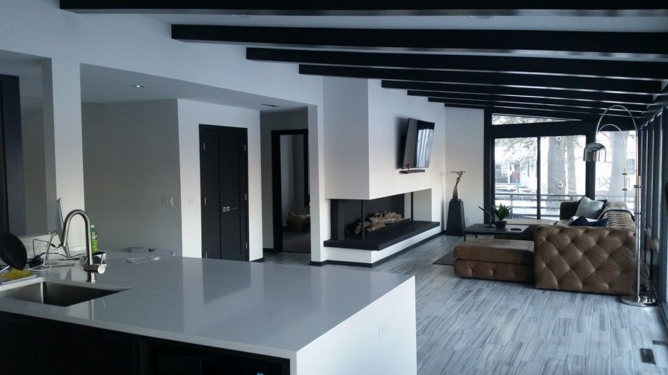 Modern living room with fireplace, kitchen island, and large windows. Black and white color scheme, wood beams.