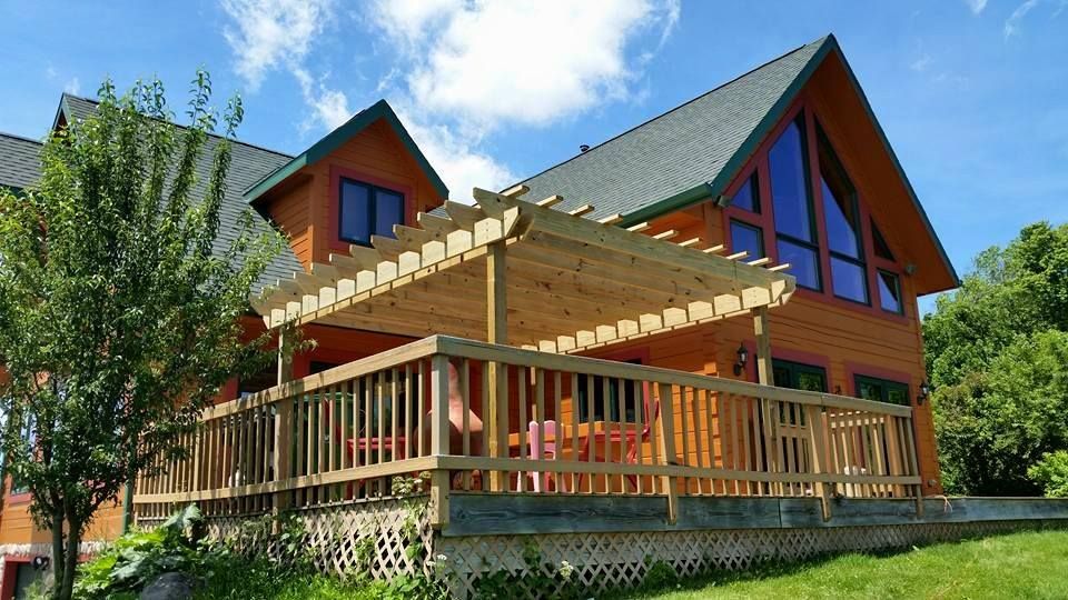 Wooden house with a deck and pergola, set against a blue sky with trees.