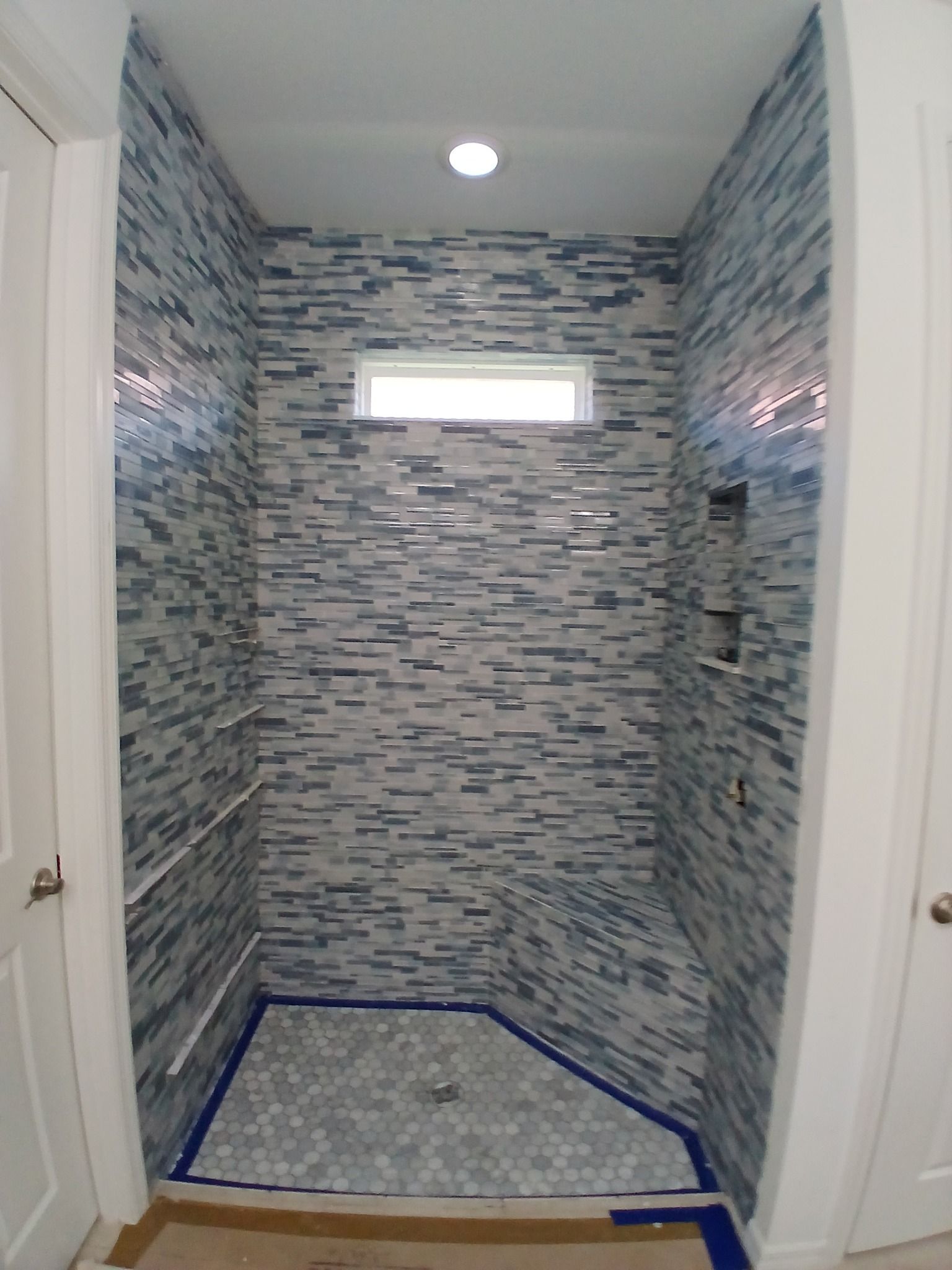 A small shower with blue and white mosaic tile walls, a small window, and a gray hexagonal tile floor.