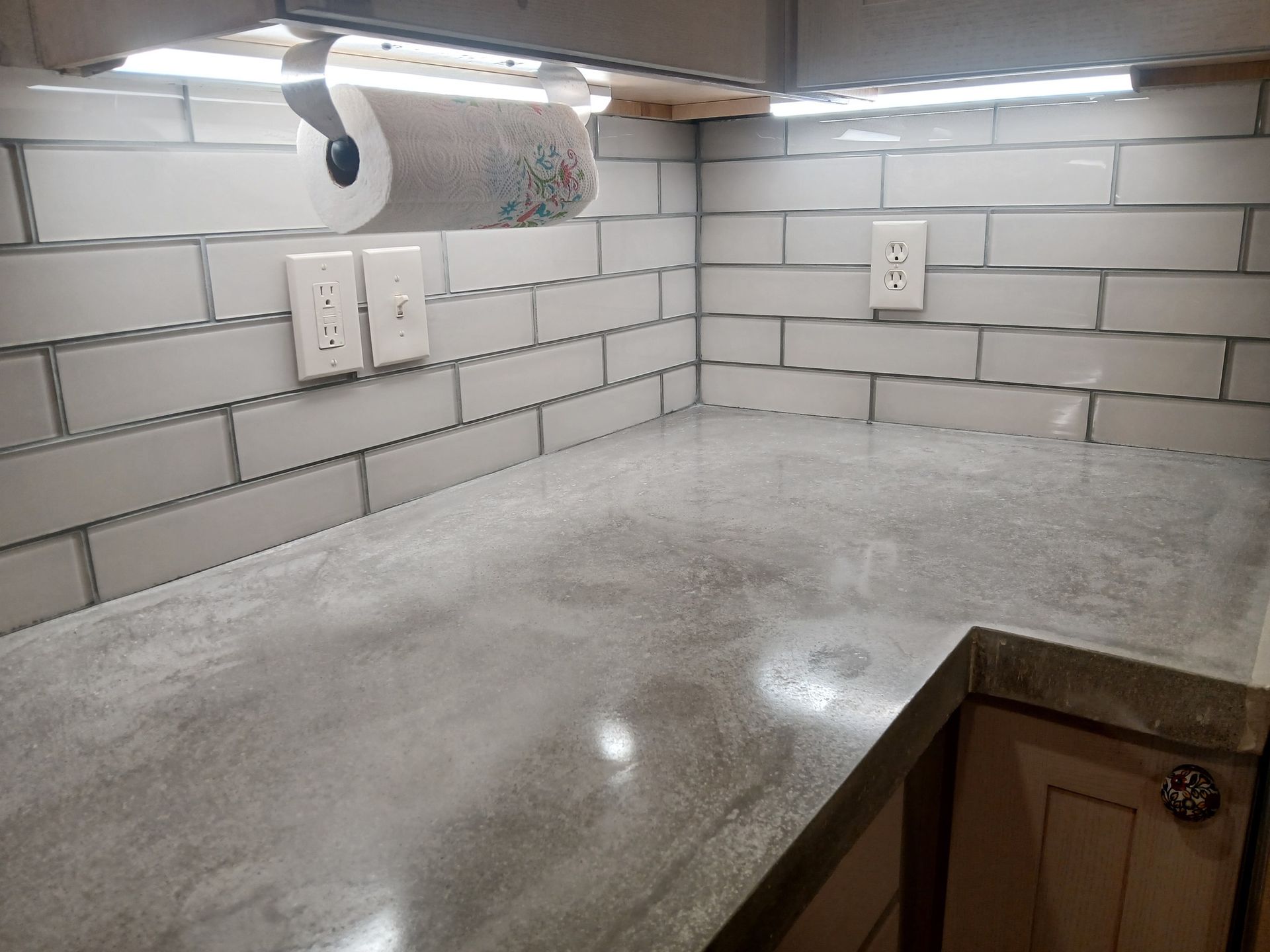 Gray kitchen countertop with white brick-style backsplash. Under-cabinet lighting and paper towel roll are visible.