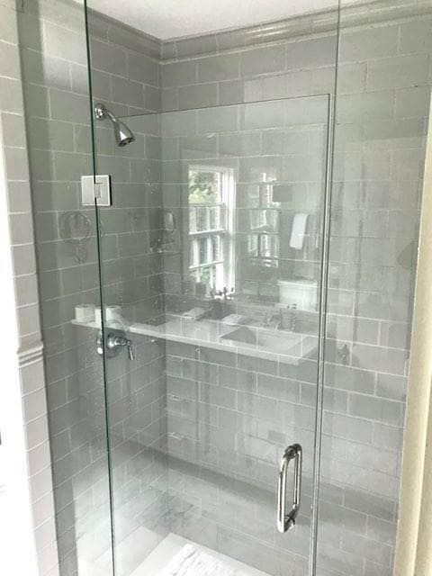Gray tiled shower with glass door and shelf, chrome fixtures, and window reflection.