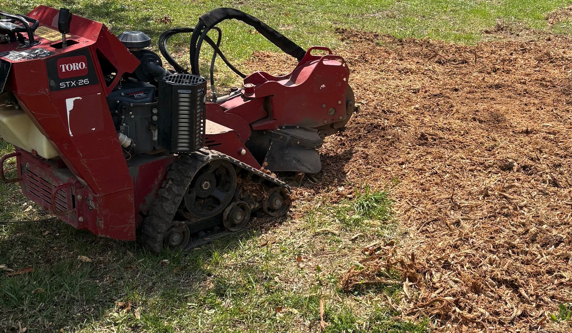 Red tracked stump grinder chewing up wood chips in a grassy yard beside a mulch-covered area