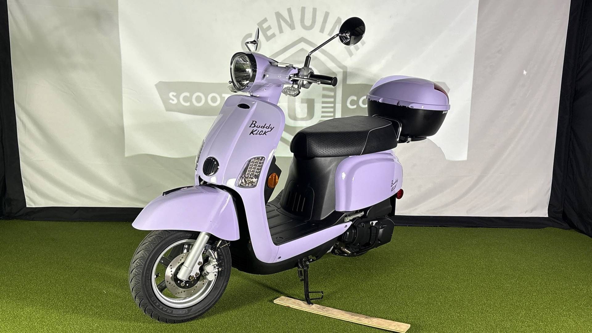A light purple Buddy scooter with a matching top case stands on green turf in front of a white branded backdrop.