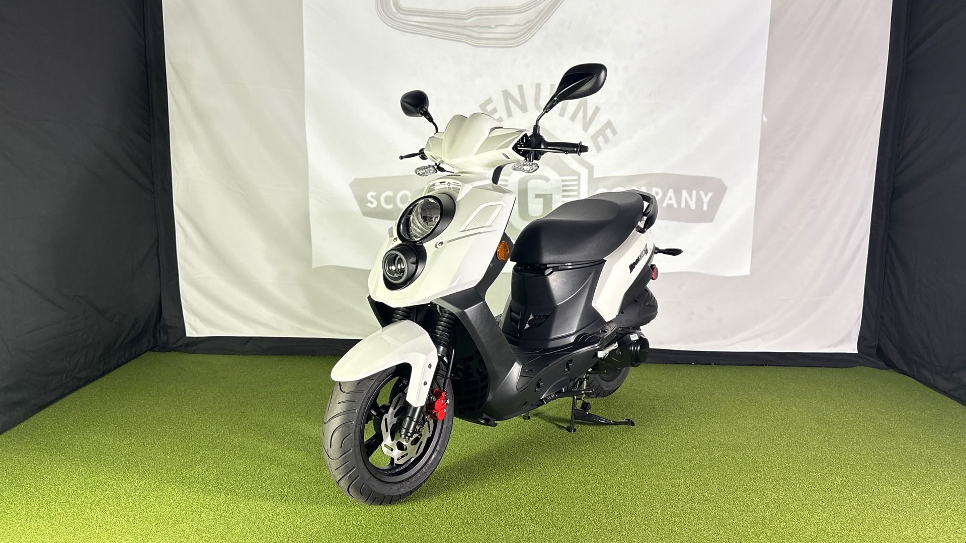 A white Genuine Scooter Company motor scooter parked on a patch of artificial grass in front of a branded backdrop.