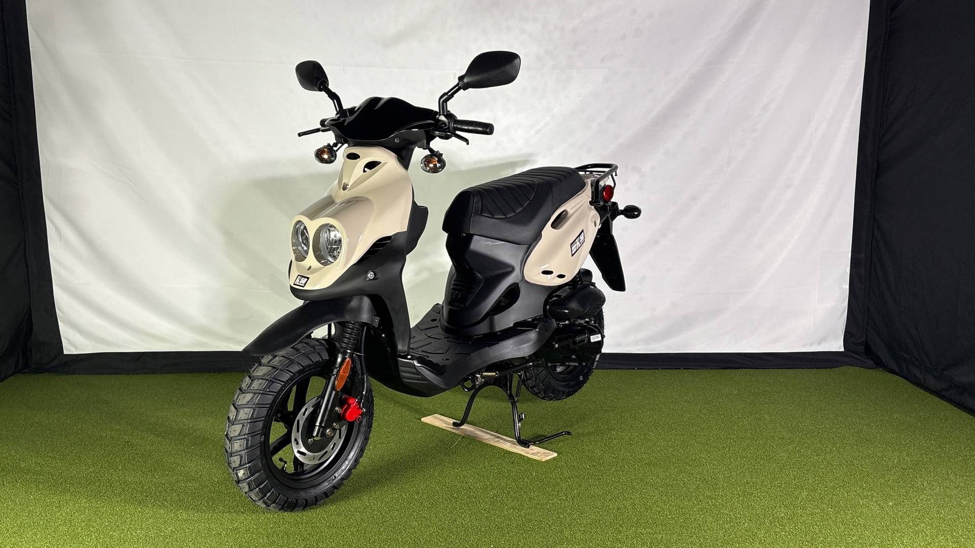 A tan and black dual-headlight scooter standing on artificial grass in a studio with a white backdrop.