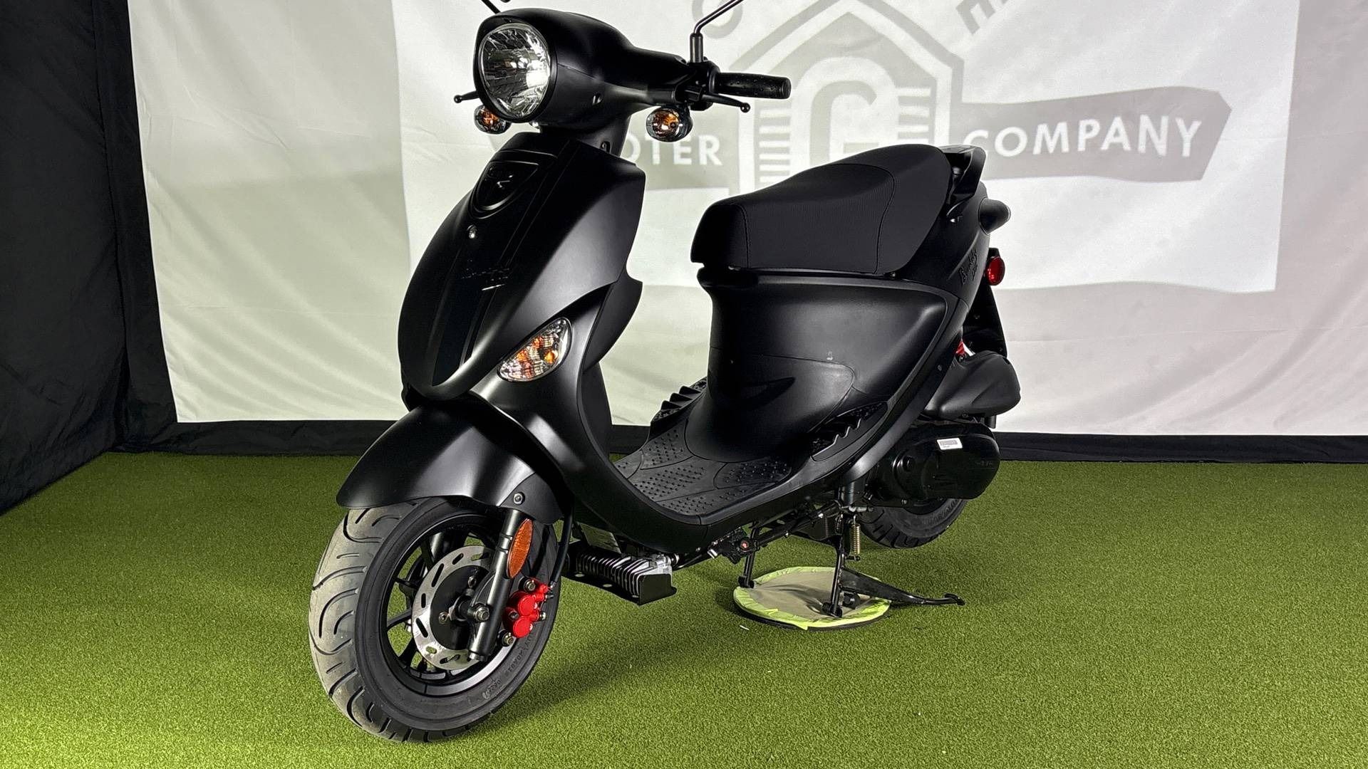 A matte black scooter stands on green turf in front of a white backdrop with a faint logo.