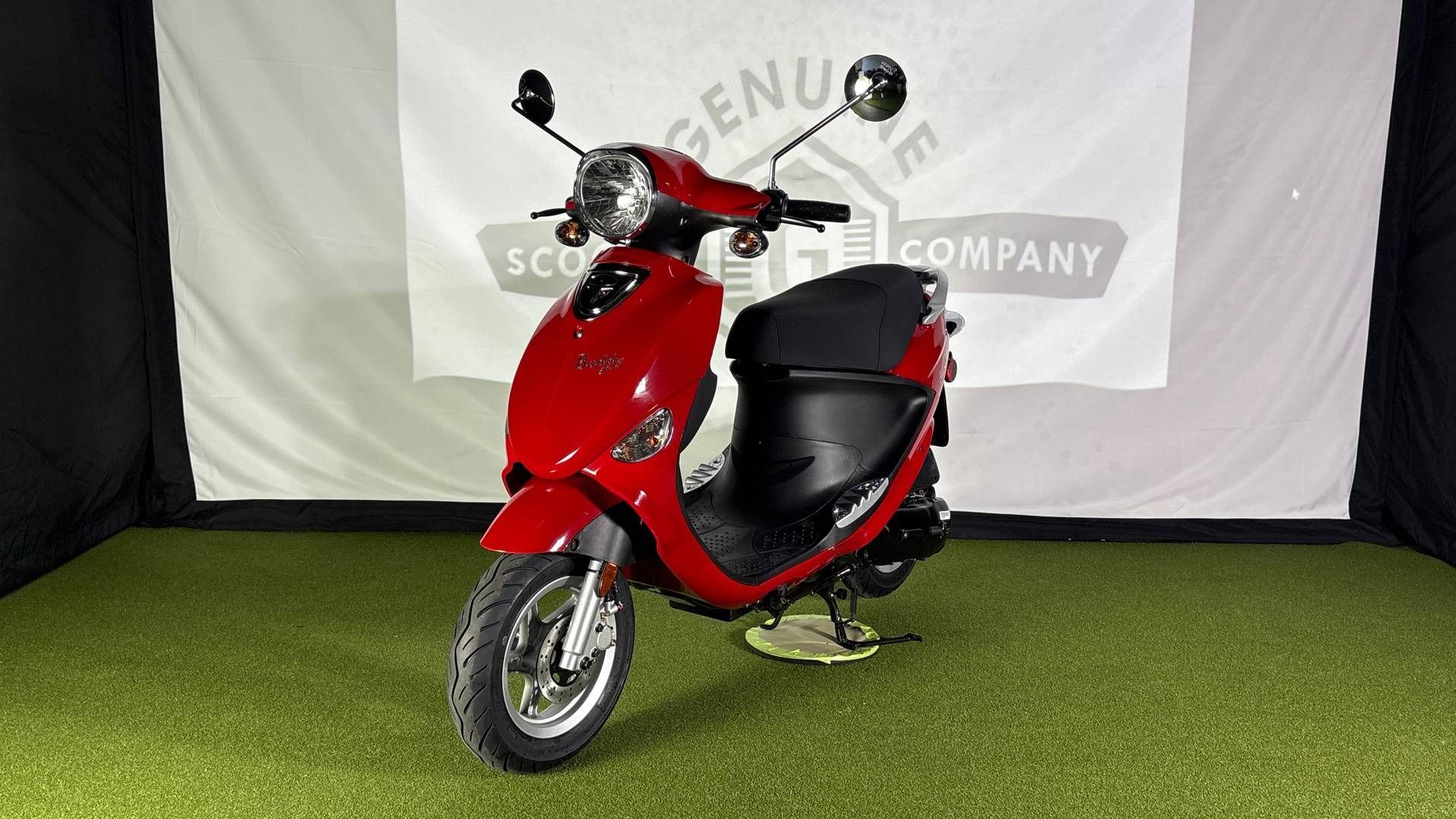 A shiny red motor scooter centered on green artificial turf against a white background featuring a logo.