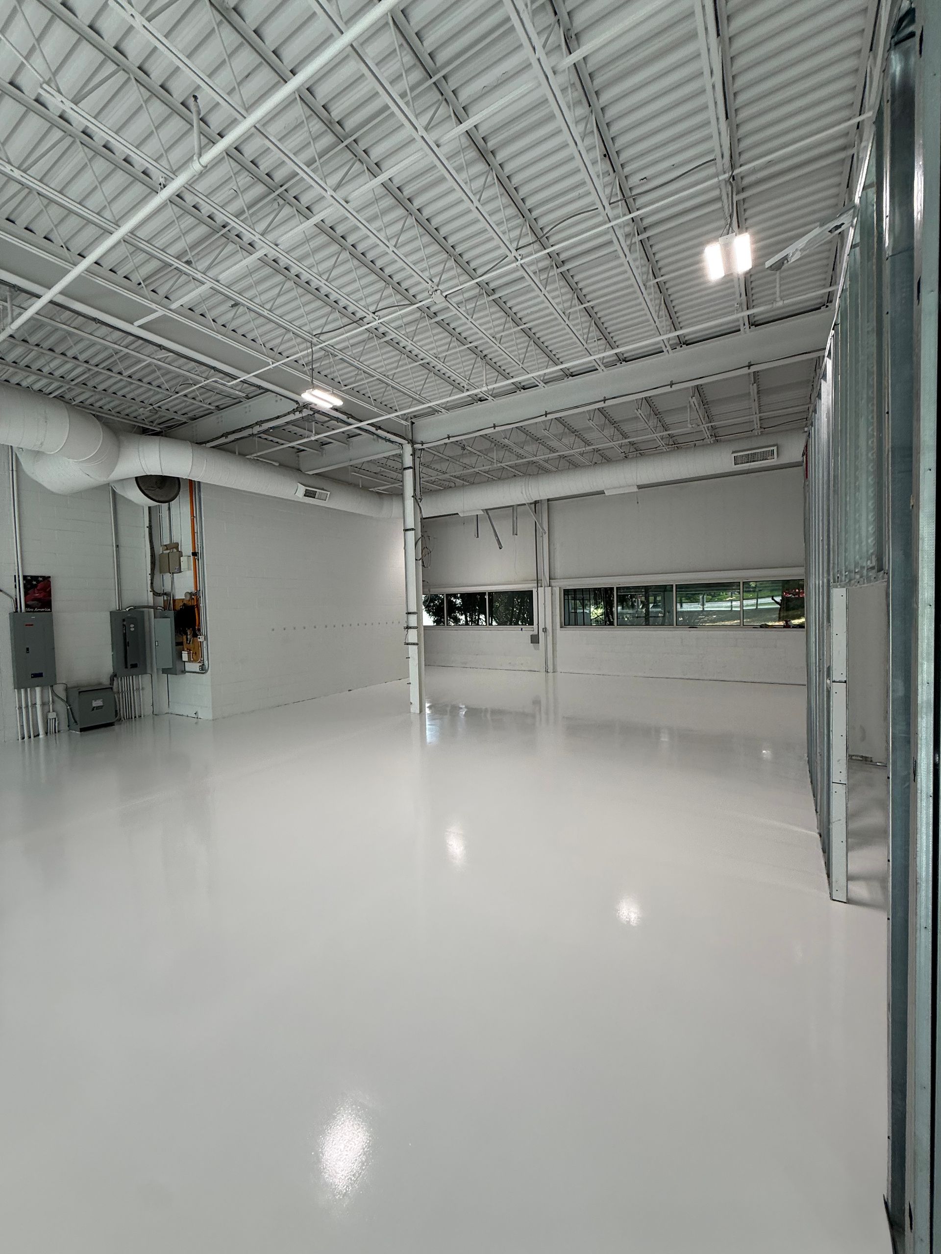Empty industrial room with glossy white floor, exposed ceiling, and large windows along one wall