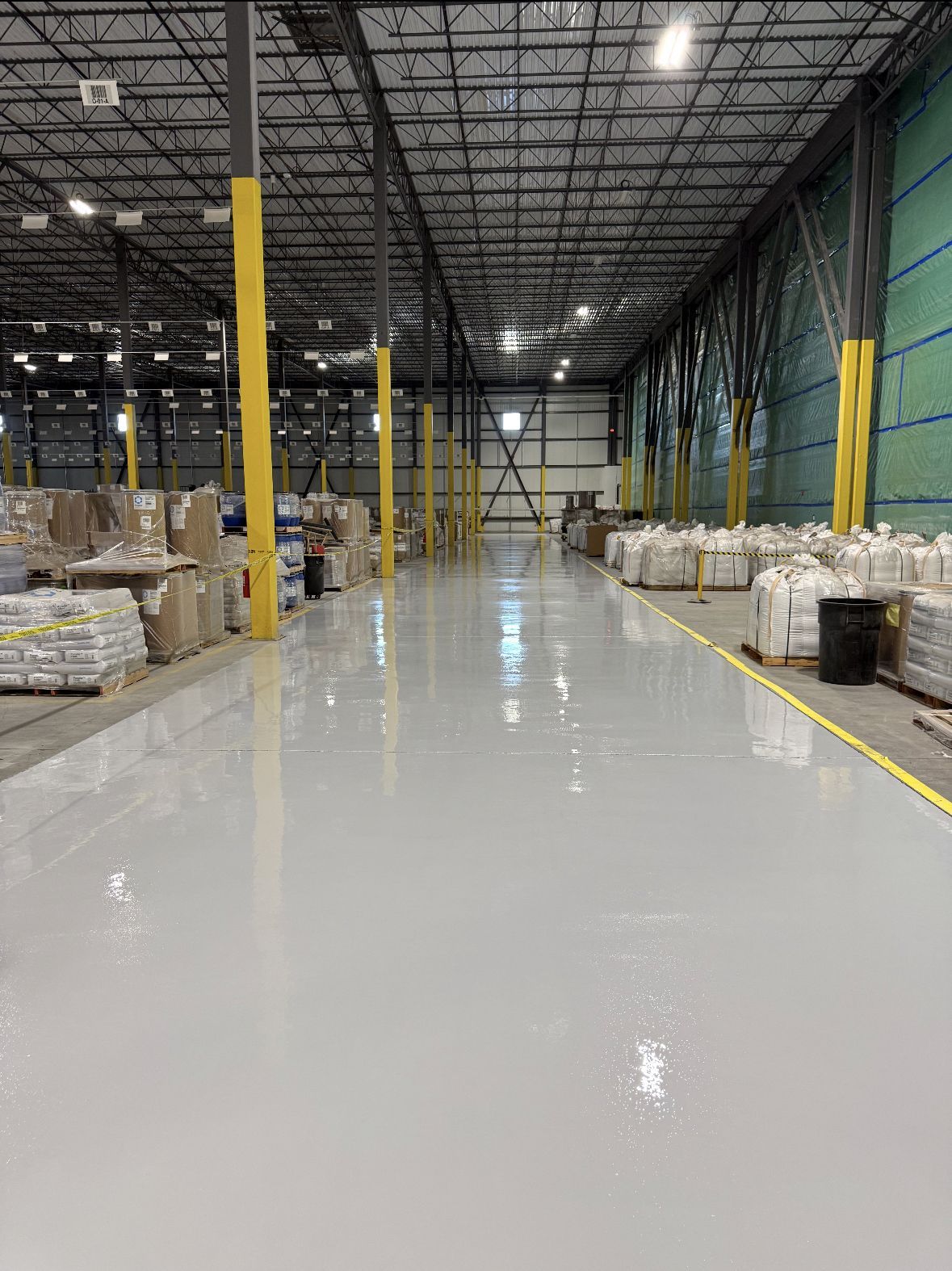 Empty warehouse aisle with glossy white floor, yellow safety posts, and stacked pallets along the sides