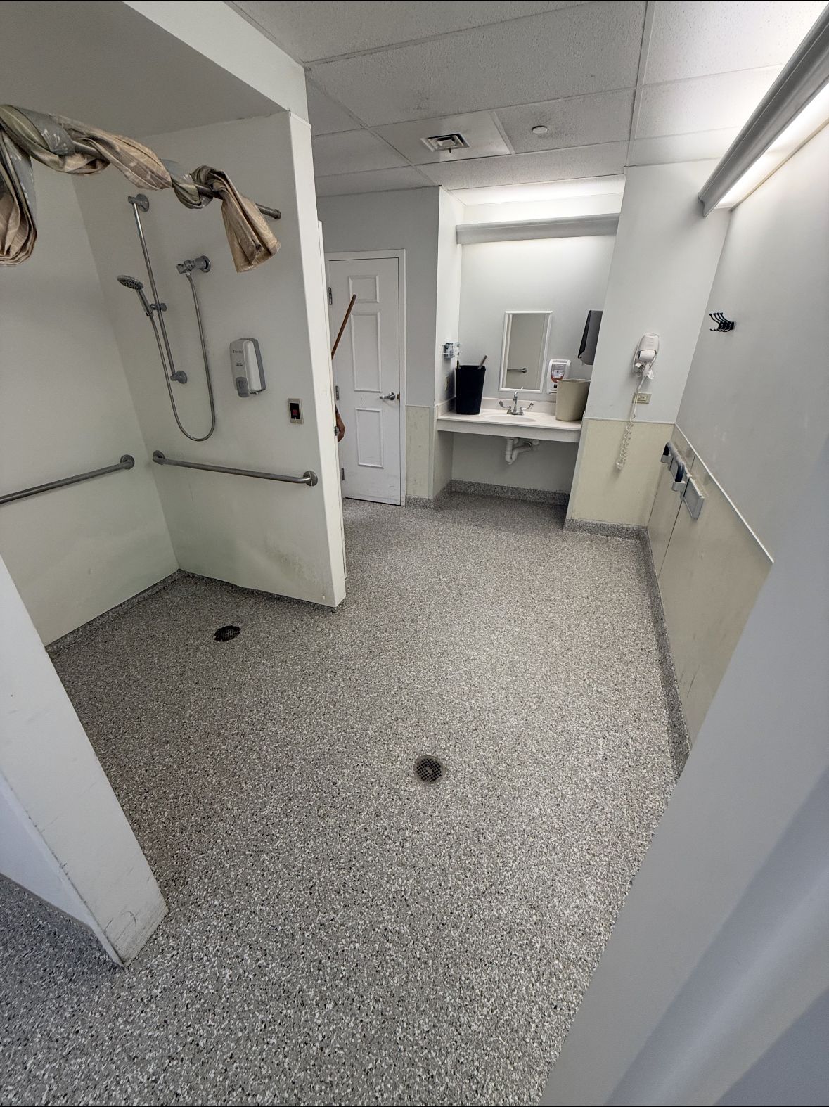 Empty white bathroom with open door, speckled tile floor, sink, mirror, and shower area visible