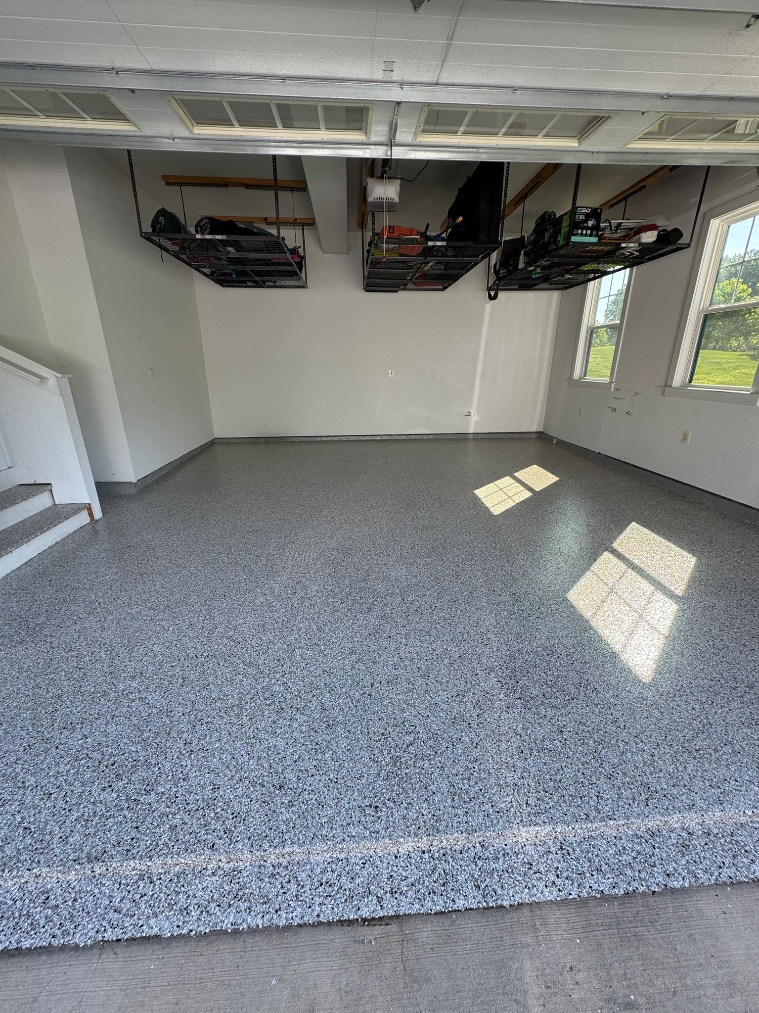 Empty garage with speckled gray floor, white walls, overhead storage racks, and sunlight from windows.
