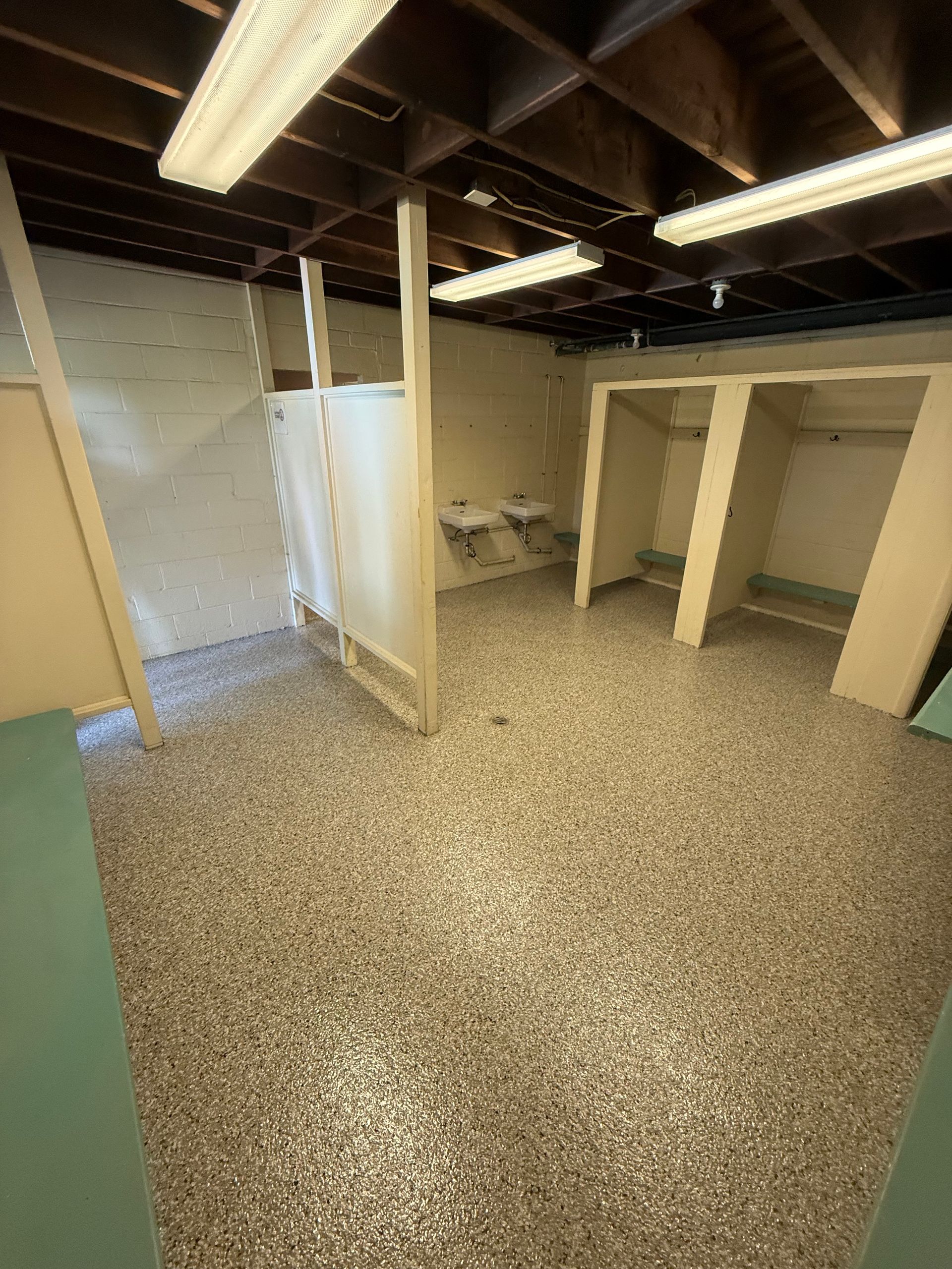 Basement restroom area with stalls, speckled floor, exposed beams, and fluorescent lights