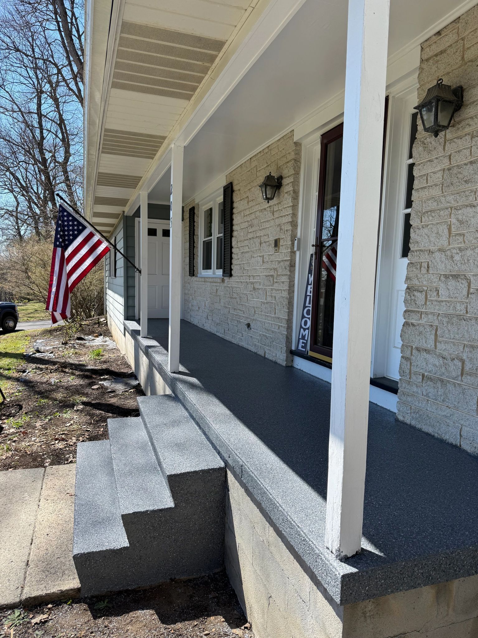 Front porch of a white brick house with a U.S. flag and columns in bright sunlight