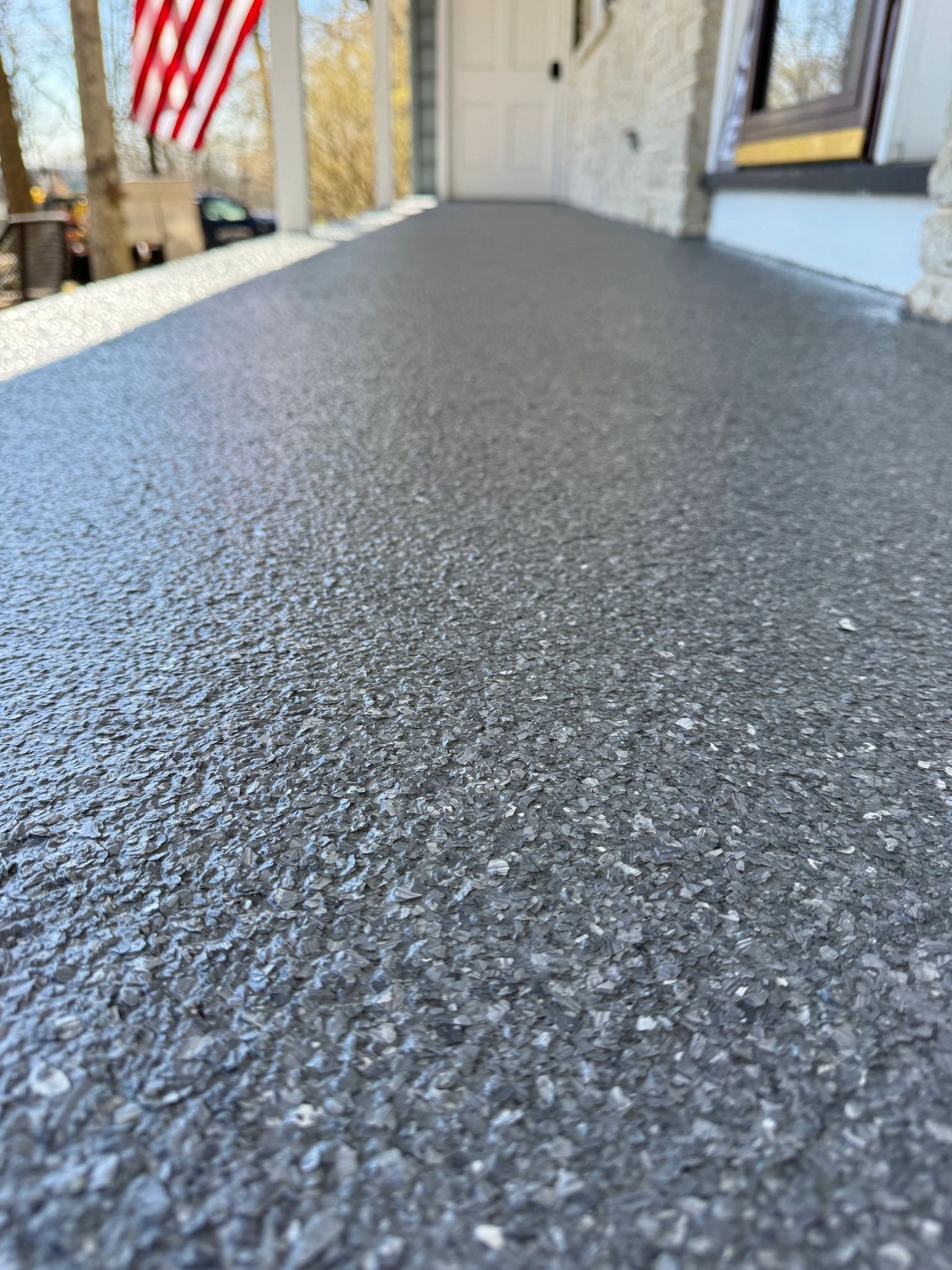 Close-up of a textured dark gray outdoor walkway beside a house with an American flag in the background