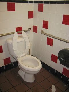 Toilet in a public restroom with white tiles, red squares, and metal grab bars.