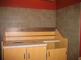 An accessible waste and recycling station with bins and wooden cabinetry. Beige tile wall and red accent.