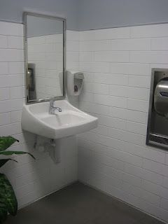 Public restroom corner with white tile walls, sink, mirror, soap dispenser, and hand dryer.