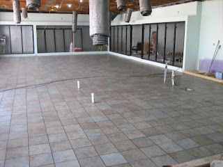 Empty room under renovation, gray tiled floor. Vertical pipes protrude. Black framed wall sections visible.