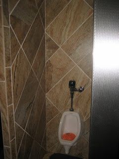 Urinal in a bathroom with tan tile and a stainless steel divider.