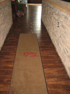 Narrow hallway with dark wood-look tile and stone walls; a brown welcome mat is centered.