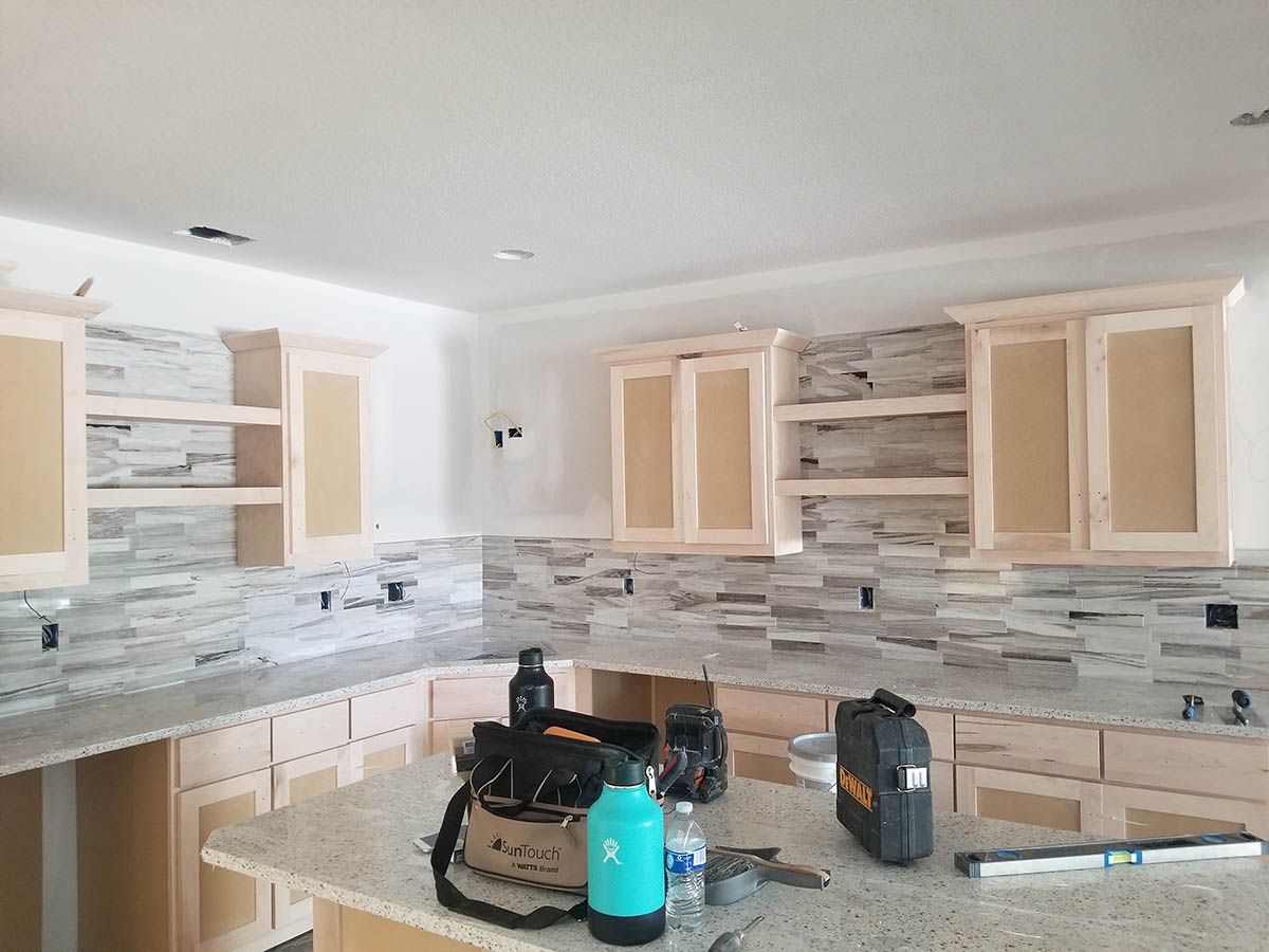 Kitchen under construction with light-colored cabinets, stone backsplash, and countertop; tools on island