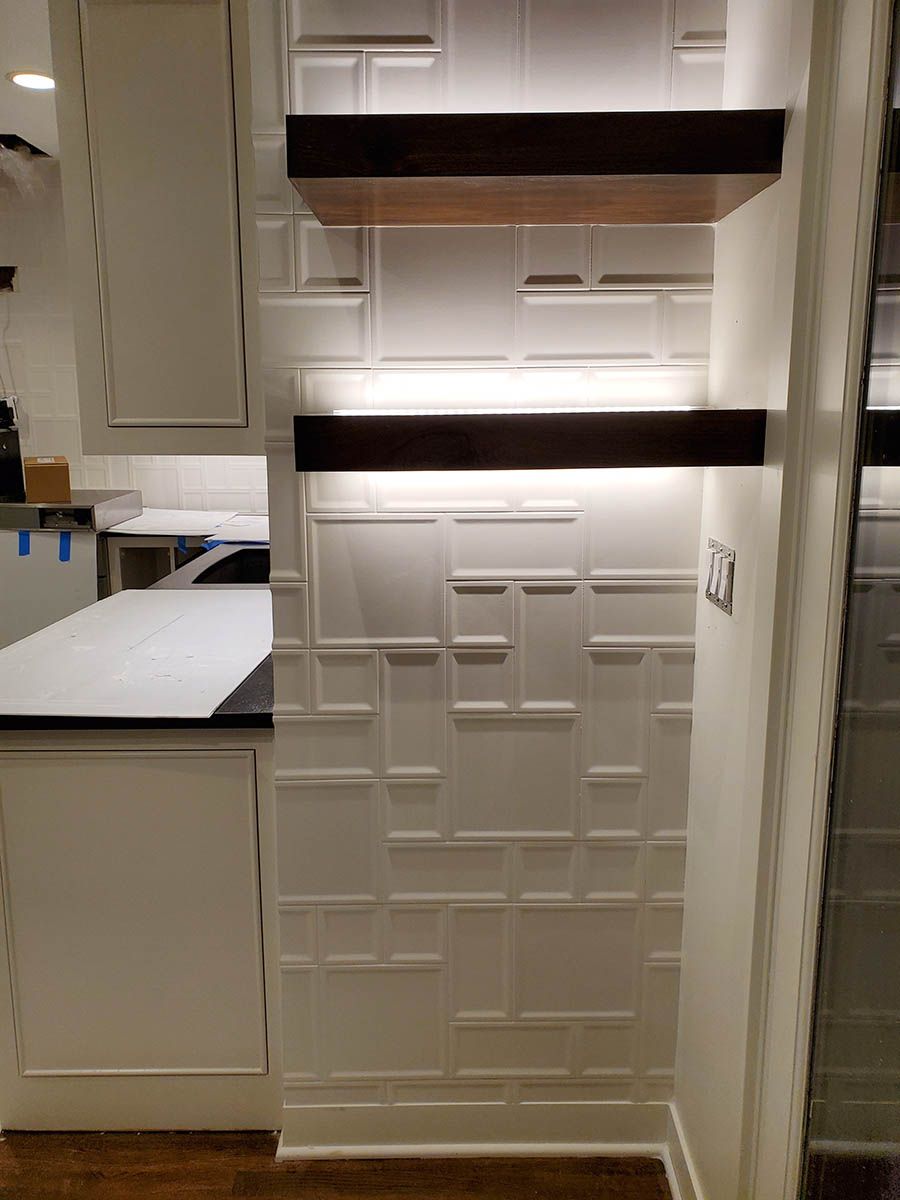 White kitchen wall with textured squares, two floating dark shelves with under-shelf lighting