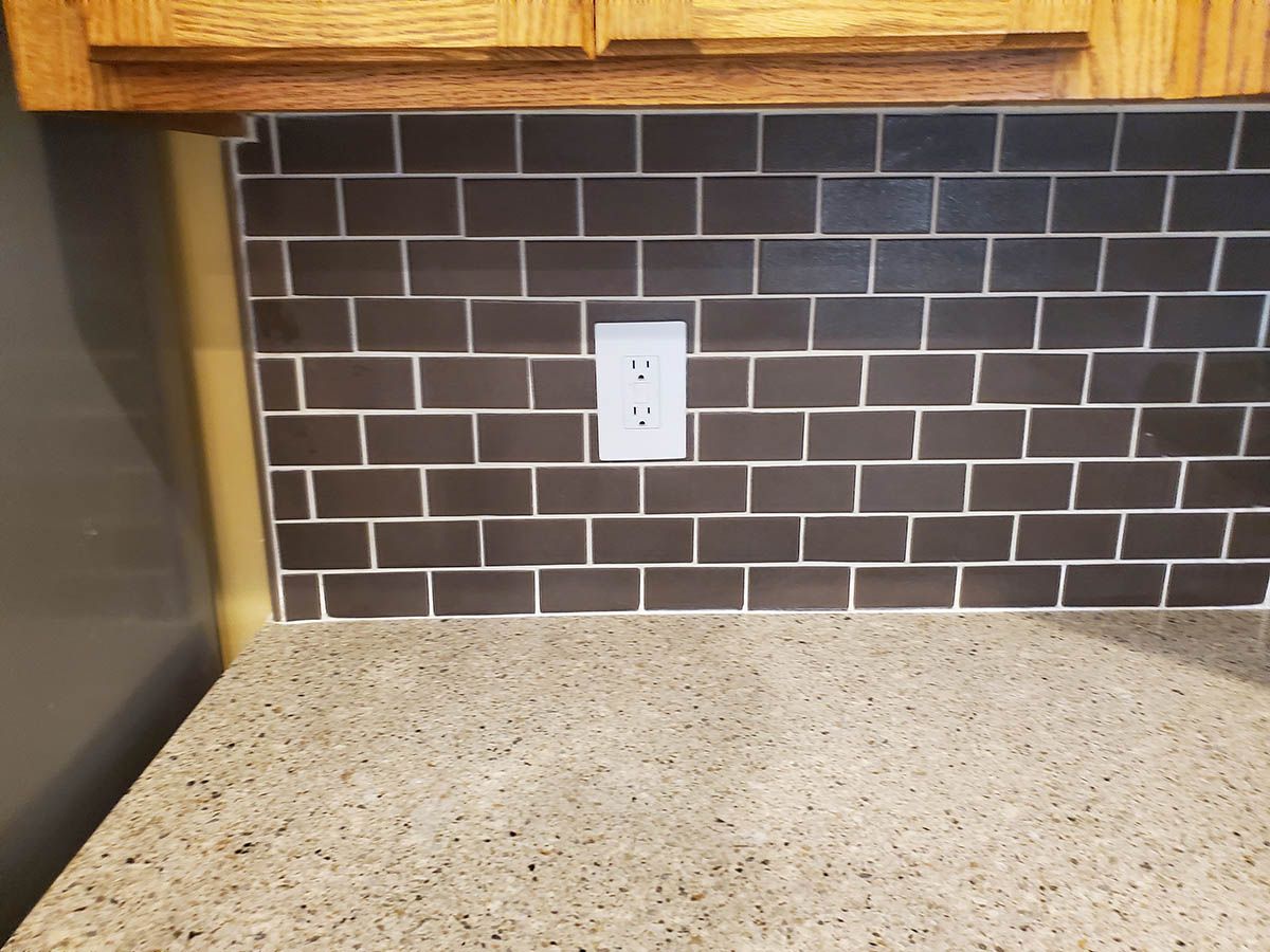 Dark brown brick-patterned tile backsplash with white grout, a white outlet, and a speckled countertop